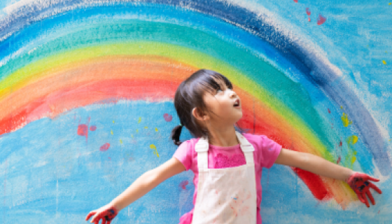 A girl stands before a rainbow painted on a wall.