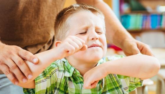 Boy of preschool age with eyes closed and bent arms up looking angry. Hands of a male educator are on his arms trying to restrain and calm him.