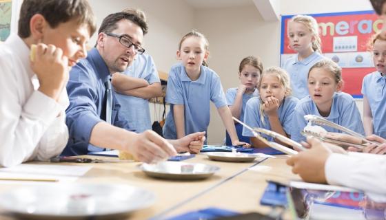 Male teacher demonstrating something to primary school children in blue uniforms, many of whom are holding tongs