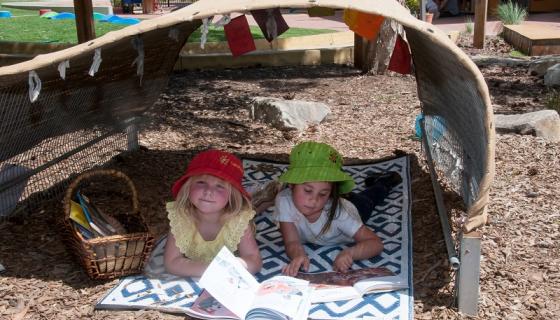 Two preschool girls wearing hats and reading on a mat outside under a shade hut